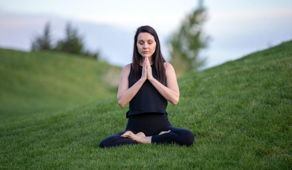 Serene woman in prayer state sitting cross legged in yoga pose outside on grassy mound
